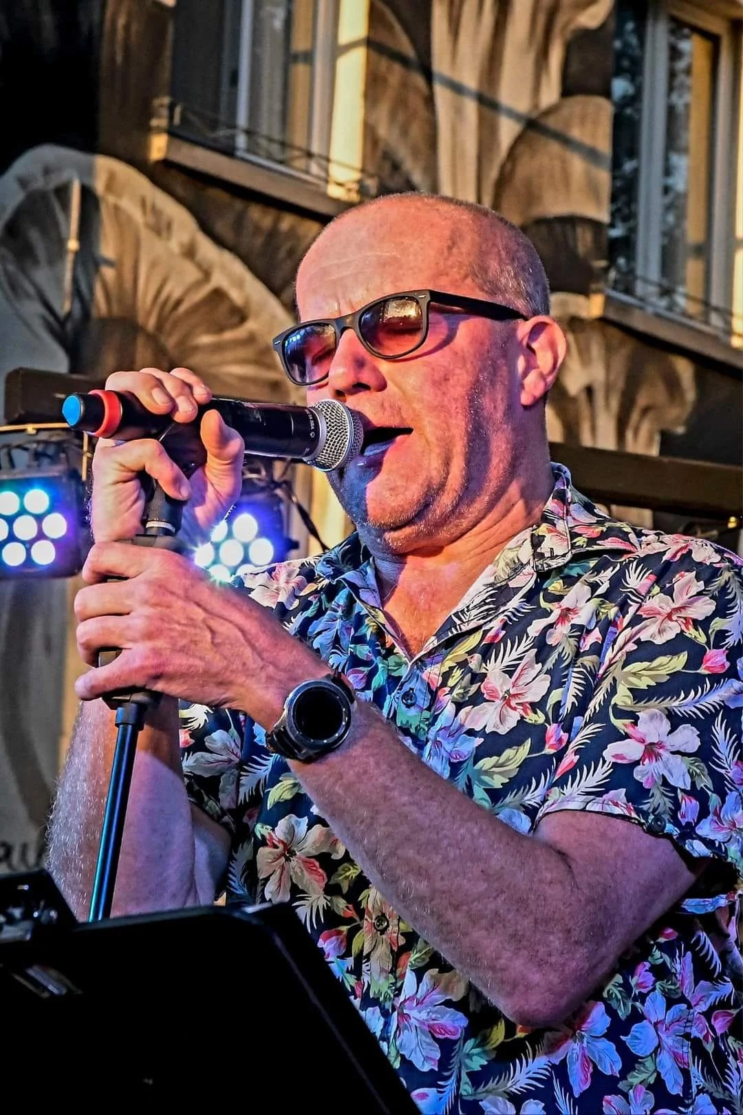 Man met een hawaii overhemd en zwart montuur zittend op een podium, zingt in een microfoon met achtergrondverlichting en een gebouw zichtbaar op de achtergrond.