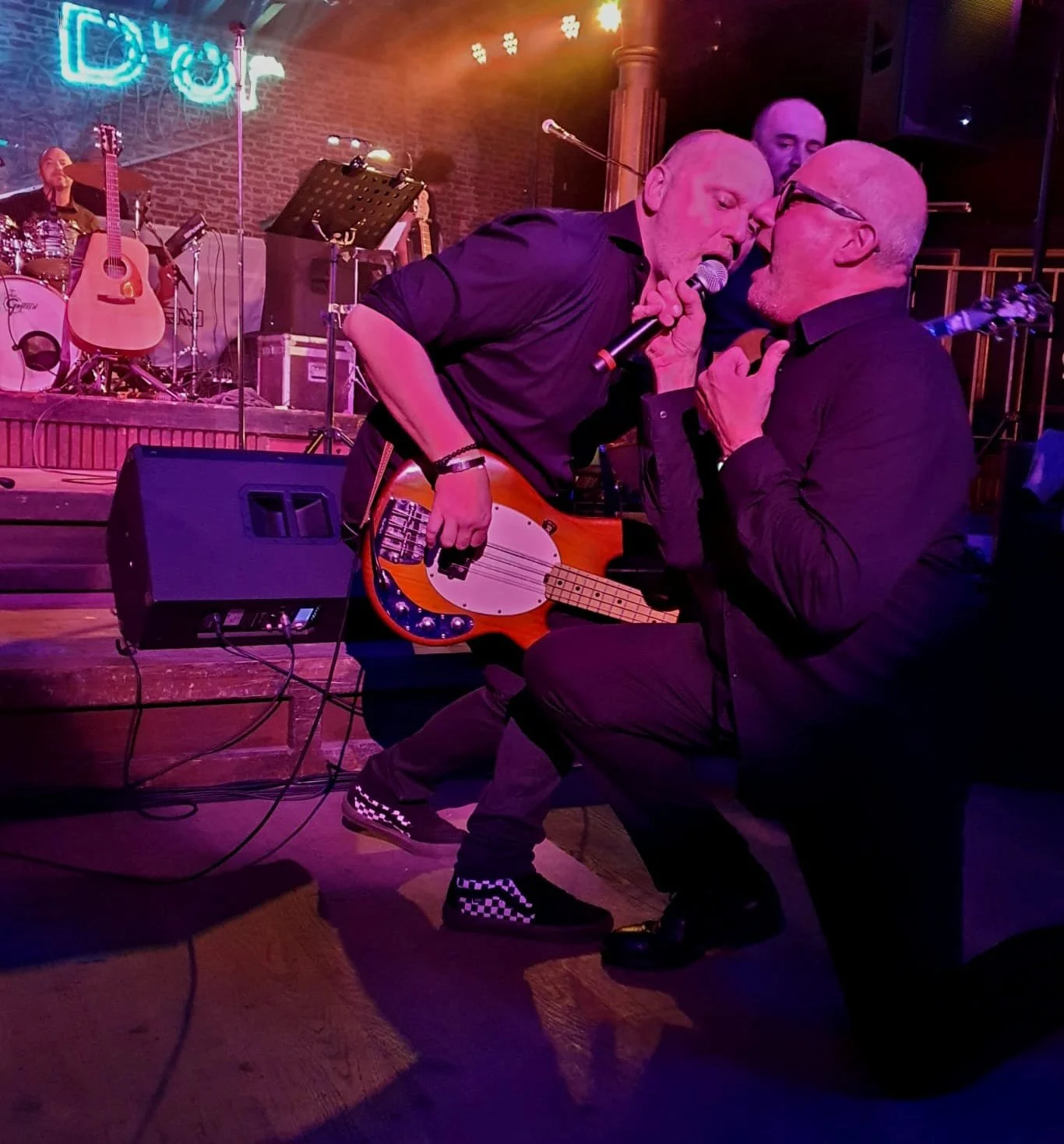 Twee mannen op het podium zingen en spelen gitaar, omringd door muziekinstrumenten en bandleden, in een club met neonverlichting.