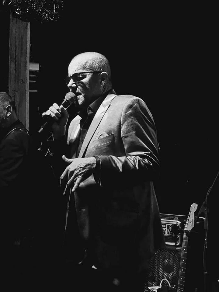 Man die in een duistere omgeving zingt of spreekt met een microfoon, gekleed in een pak en zonnebril.