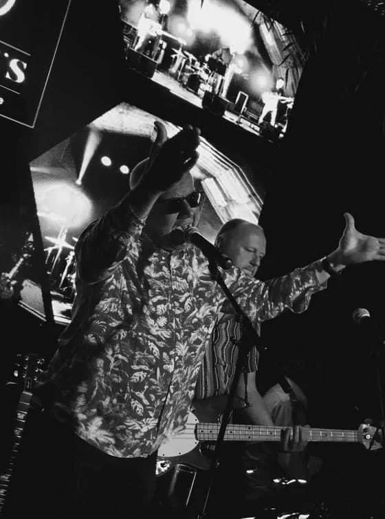 Man met microfoon en gitaar op het podium, live muziekoptreden in een club of bar, met achtergrondbeelden op schermen