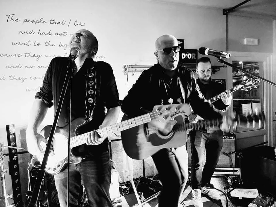 Een driepersoonlijke band speelt live muziek in een café of bar, met twee gitaristen en een zanger.