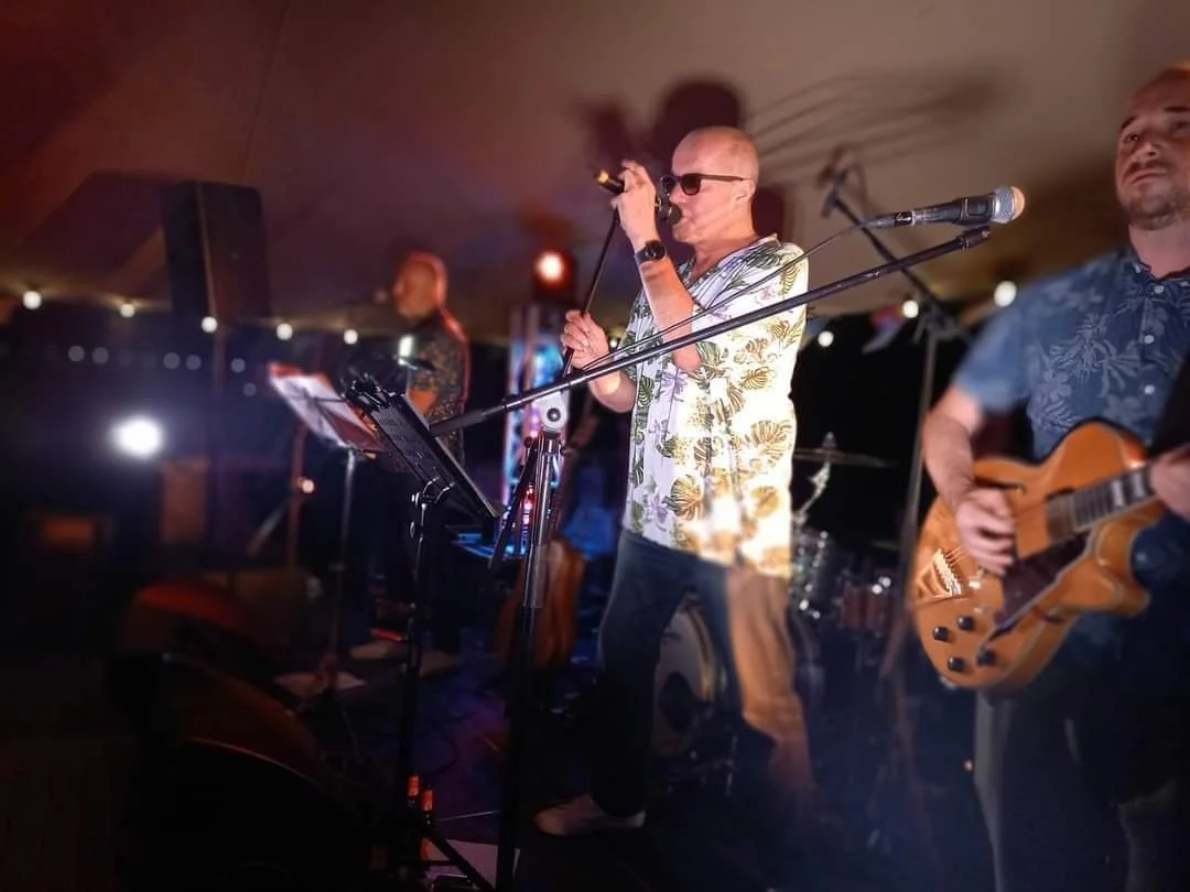 Mannen zingen en spelen gitaar en keyboard op een podium, met een microfoon, in een ontspannen sfeer.