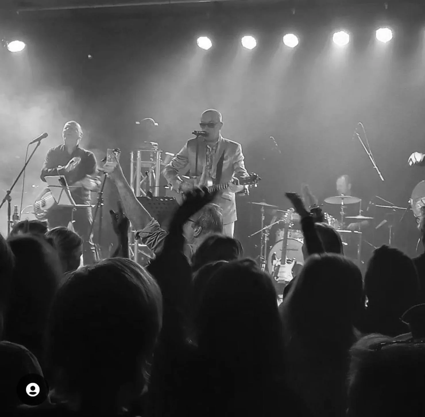 Een live muziekoptreden met een groep muzikanten op een podium, inclusief een zanger met een gitaar en een drumstel, terwijl het publiek enthousiasme uitdrukt.