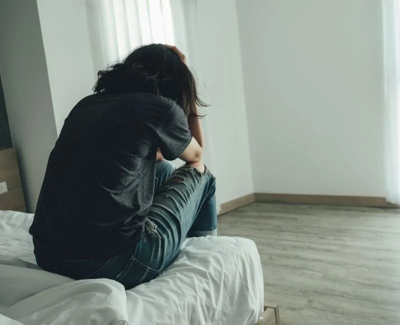 Person sitting on a bed, wearing a black shirt and ripped jeans, with their head in their hands, appearing distressed.