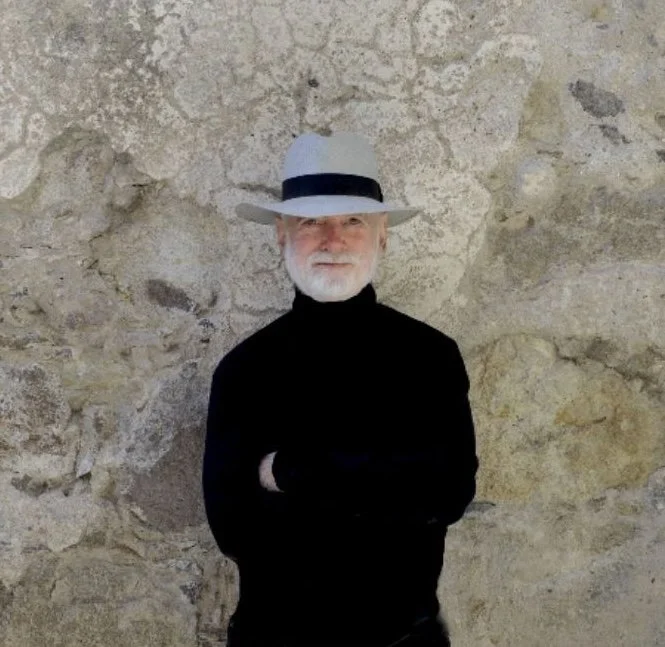 Older man with white beard in black turtleneck and white fedora hat standing against a stone wall with arms crossed.