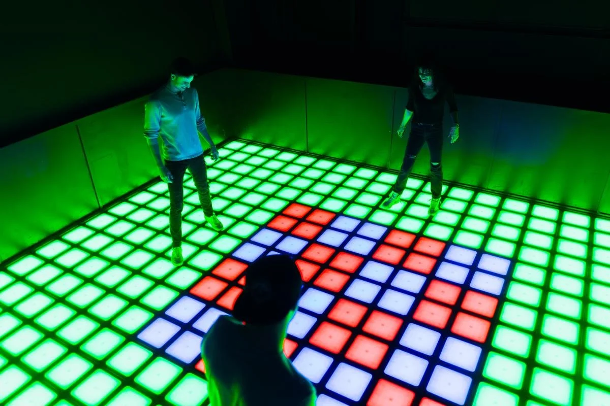 Three people standing on an illuminated dance floor with red, blue, and green LED tiles, featuring a pixelated heart pattern.