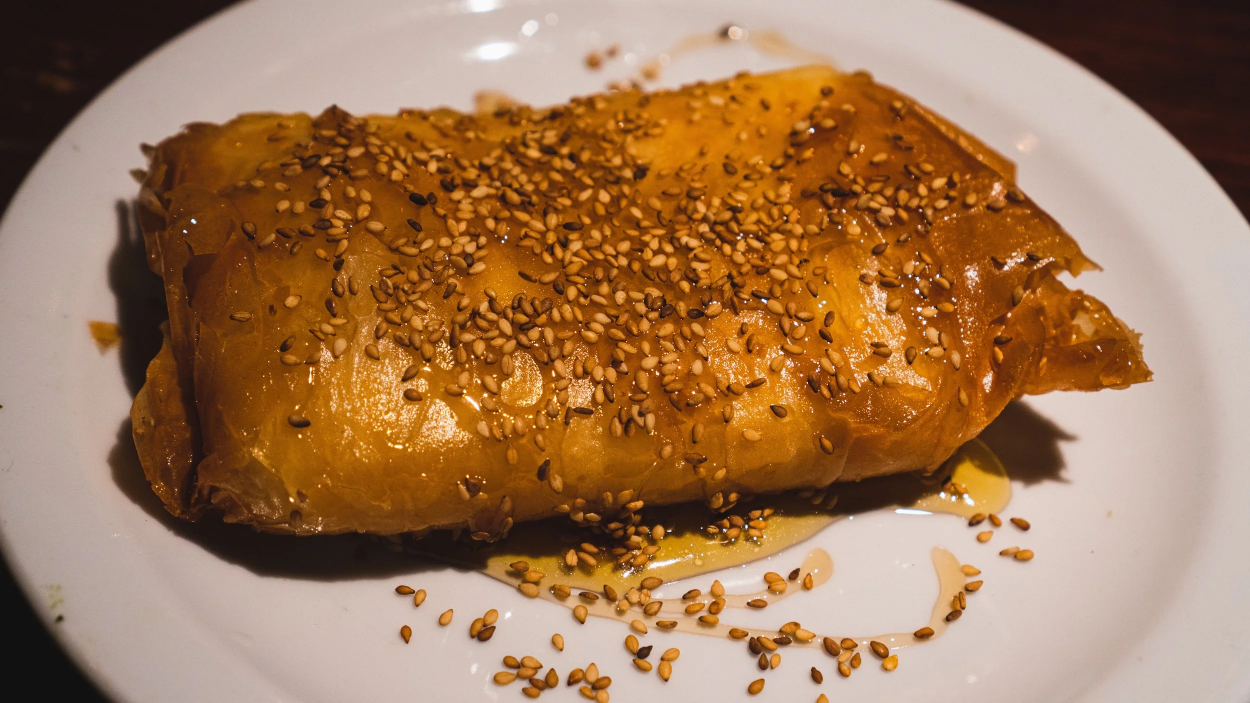 A plate with a deep fried feta cheese pastry topped with sesame seeds, drizzled with honey