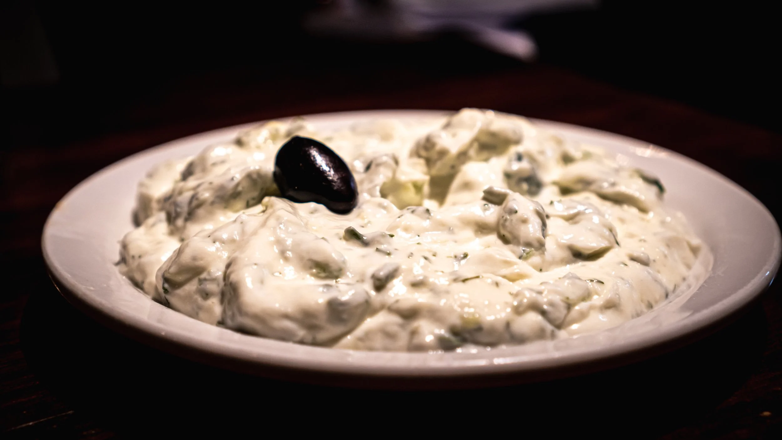 A white plate filled with creamy Greek-style tzatziki sauce containing chopped cucumbers, herbs, and a black olive on top.