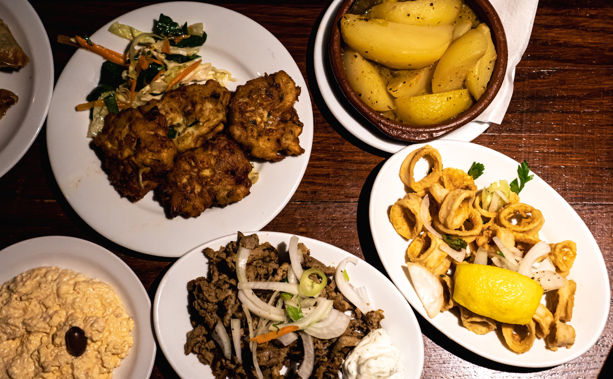 An assortment of Greek dishes on a wooden table, including fried cauliflower with a side of mixed salad, cooked potatoes, fried calamari with lemon, tabbouleh, tirosalata topped with a black olive.