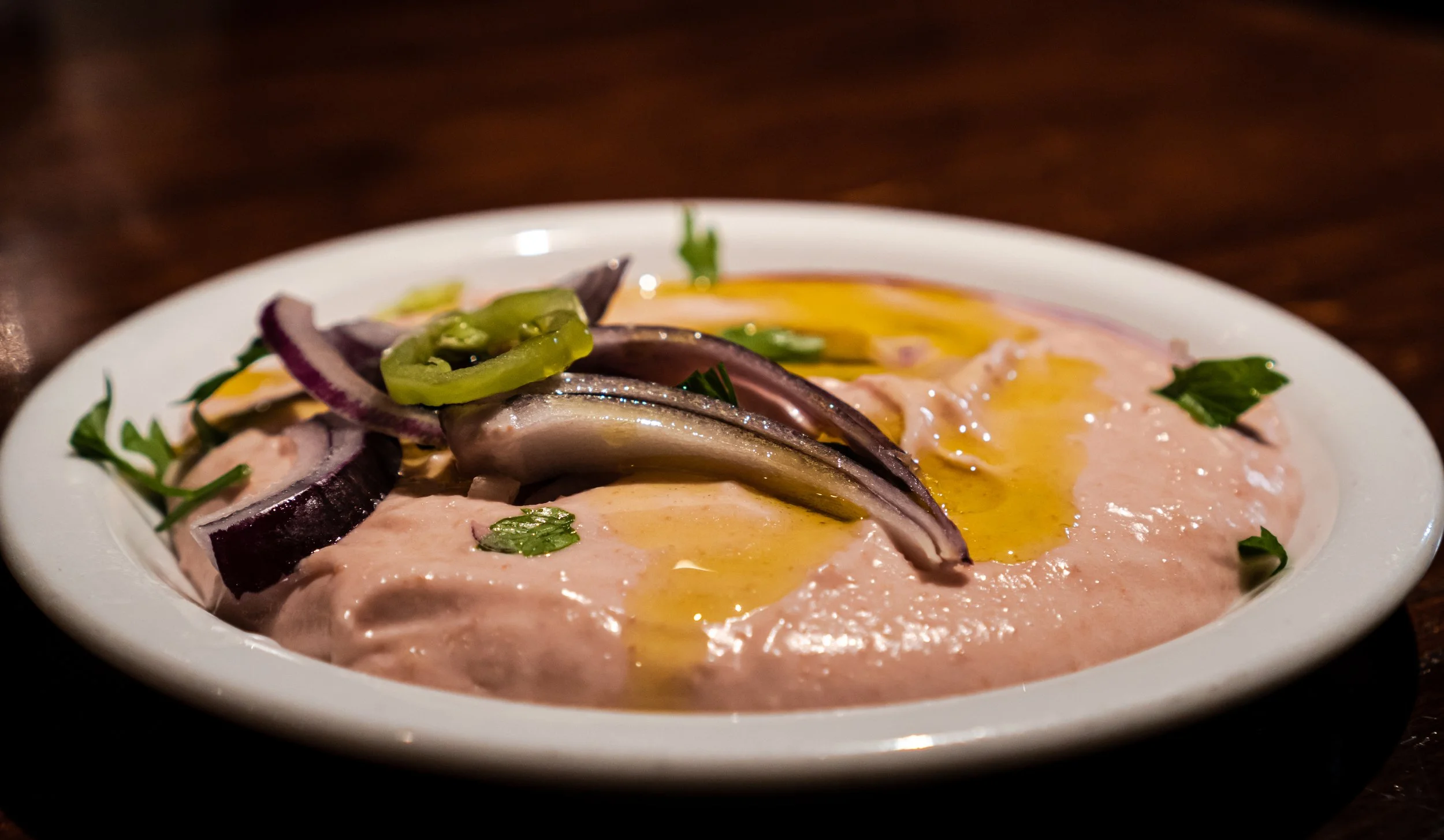 A white bowl filled with a creamy pinkish tarama topped with sliced red onions, green pepper slices, a drizzle of olive oil, and garnished with chopped parsley.