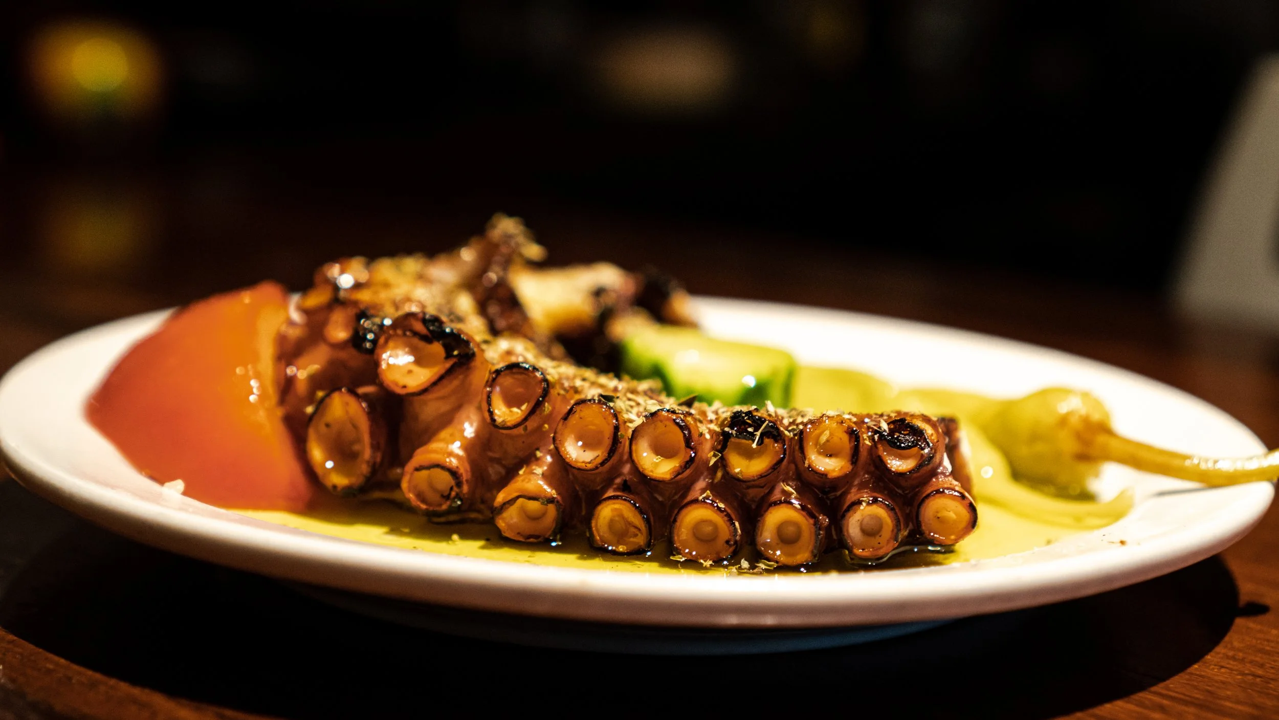 Grilled octopus tentacle served on a white plate with some vegetables and sauce.