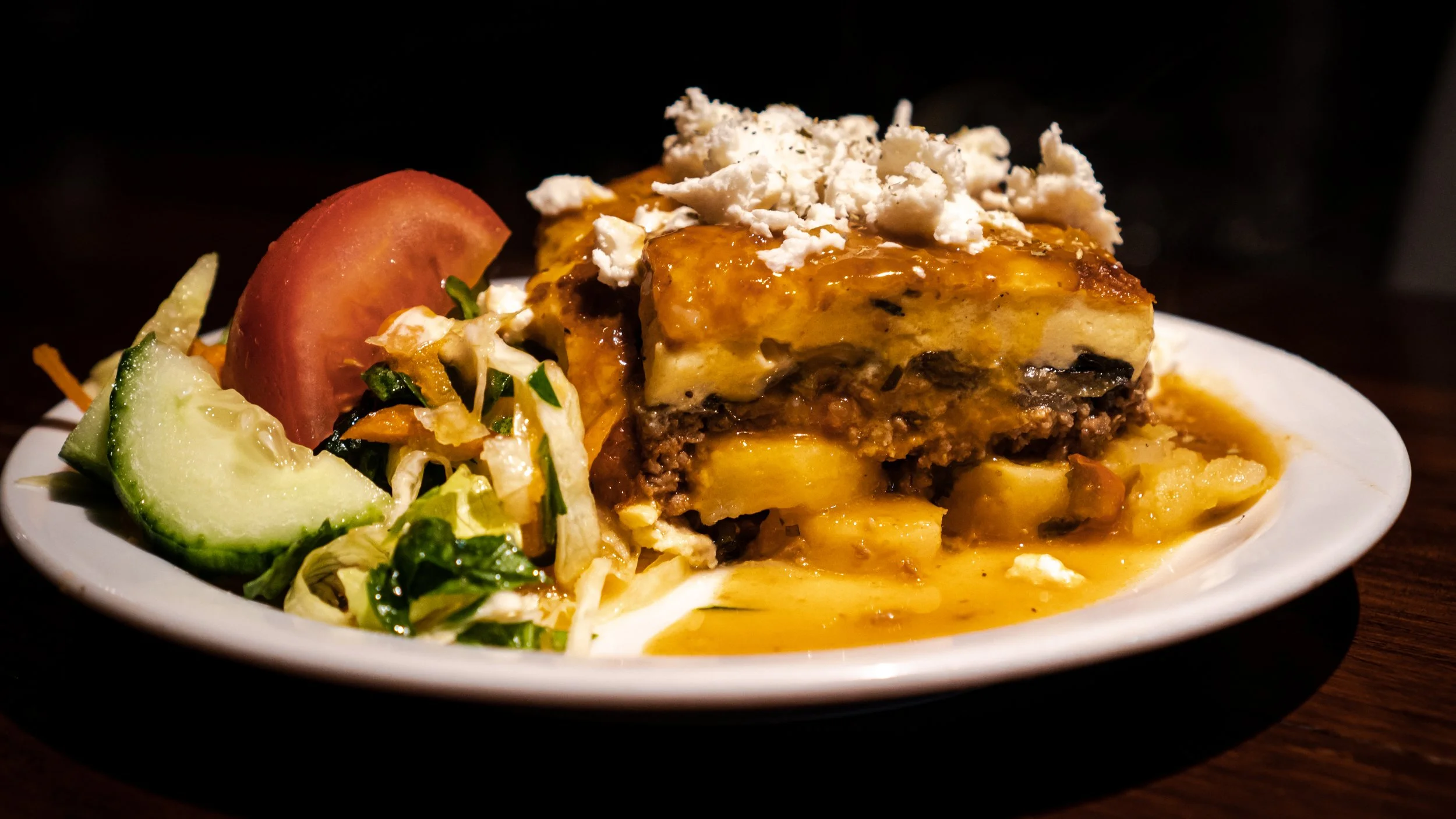 Plate of layered Mousaka with cheese, ground meat, and potatoes, topped with crumbled cheese, served with a salad of lettuce, cucumber, and tomato.