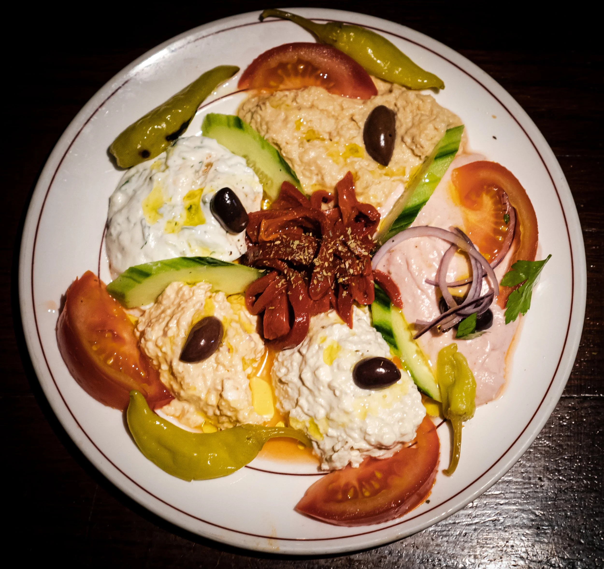 A plate of assorted Mediterranean appetizers including tomato slices, cucumber, green chili peppers, hummus, tzatziki, sliced meats, olives, onions, and parsley.