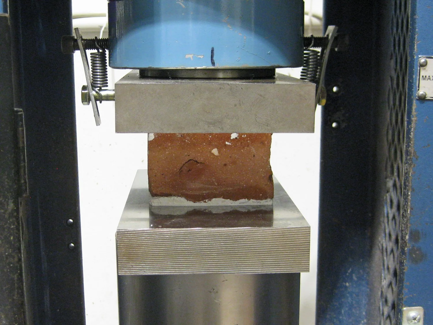 A gypsum-capped red brick specimen loaded between hardened steel platens in a compressive strength machine.