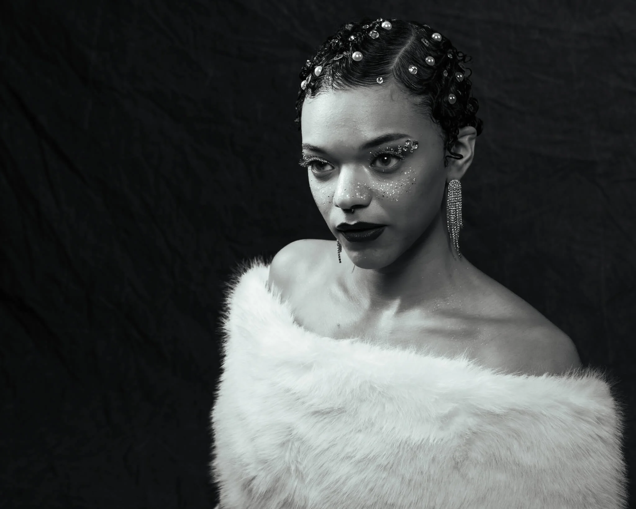 lack and white portrait of a woman in an off-shoulder white fur wrap gazing intensely at the camera, wearing long crystal fringe earrings, glitter eye makeup, glitter freckles, dark lips, and pearl and gem hair gems against a dark draped backdrop