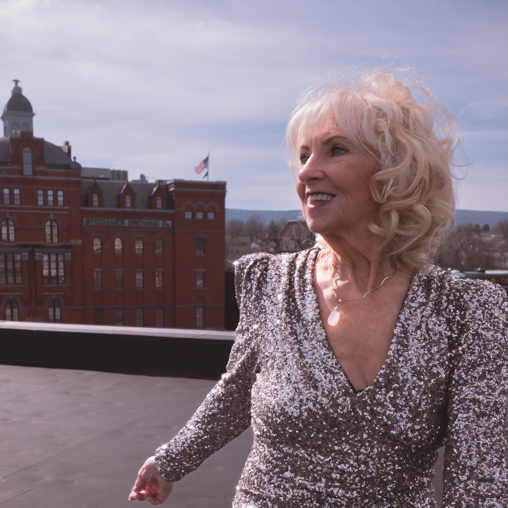Rooftop portrait session above downtown Wilkes-Barre, Pennsylvania, with the city's historic architecture visible in the background. A woman in a sequined gown catches natural light against an open sky — bold, radiant, and completely herself. Paulett
