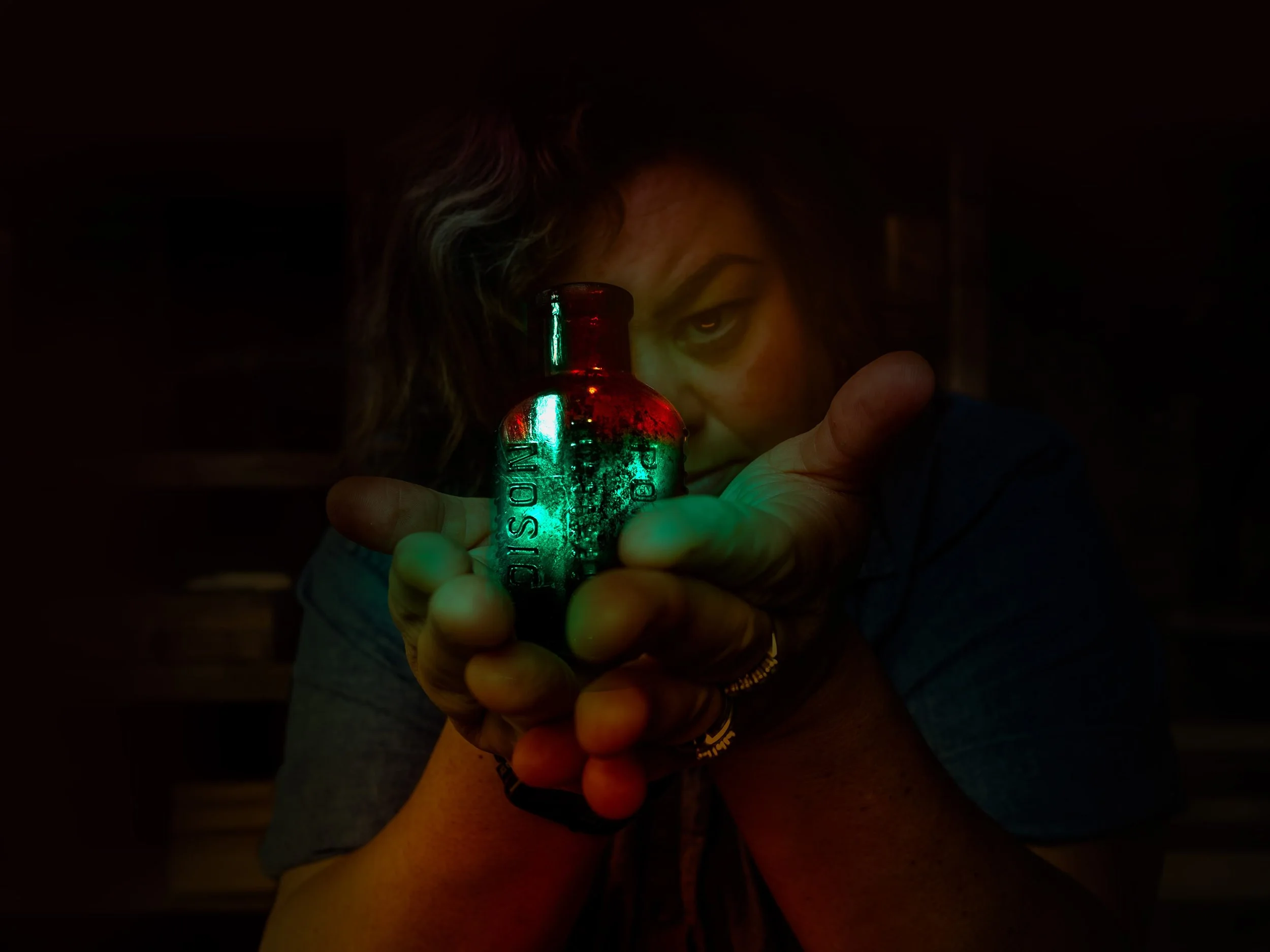 Dark self-portrait photograph of a woman extending an antique glass POISON bottle toward the camera, one eye visible through her hair, bioluminescent green and amber light glowing from within the bottle, fine art photography by Paulette Michelle.