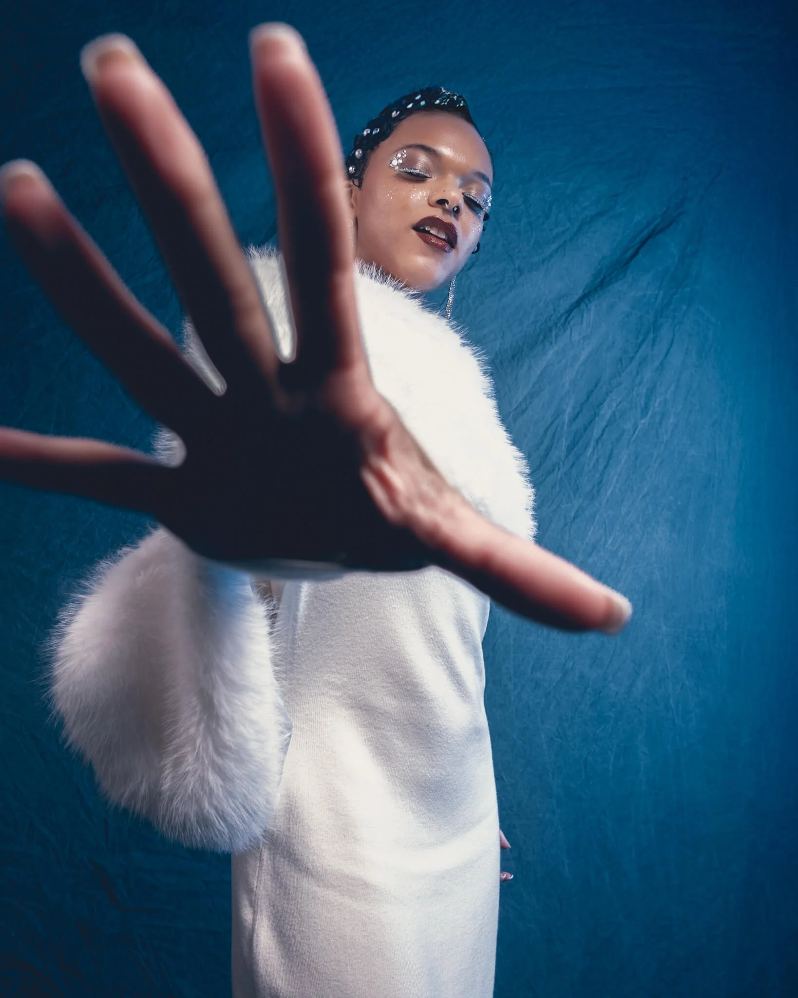 Woman in a white dress and fur stole extends one hand toward the camera in a blurred stop gesture, eyes closed, wearing glitter eye makeup and crystal hair gems against a deep blue textured backdrop