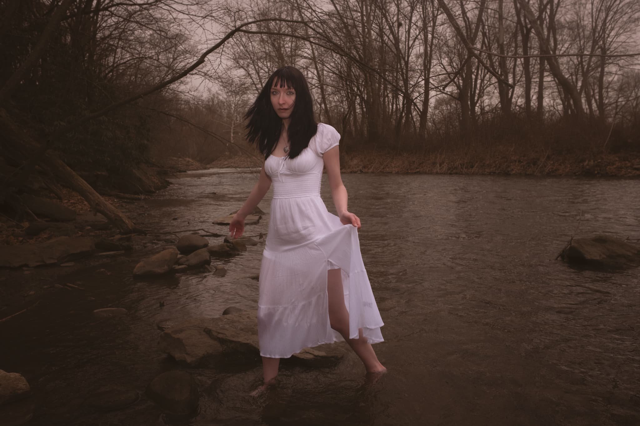 Fine art portrait of a woman in a white dress standing barefoot in a creek bed, surrounded by bare winter trees in Scranton, Pennsylvania. Equal parts haunting and alive — this is the kind of portrait that lives somewhere between folklore and memory.