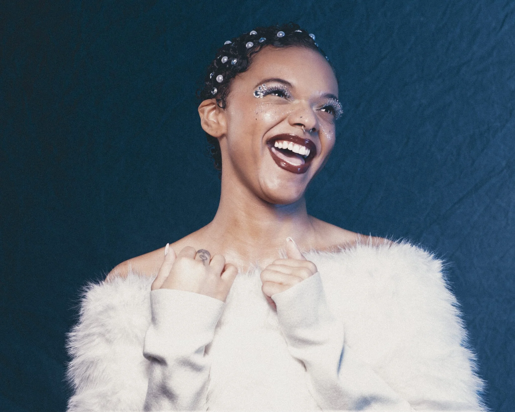 Woman laughing with head tilted back, both hands gripping the lapels of a white fur stole, wearing glitter eye makeup, silver freckles, dark red lips, and pearl hair gems against a dark teal backdrop