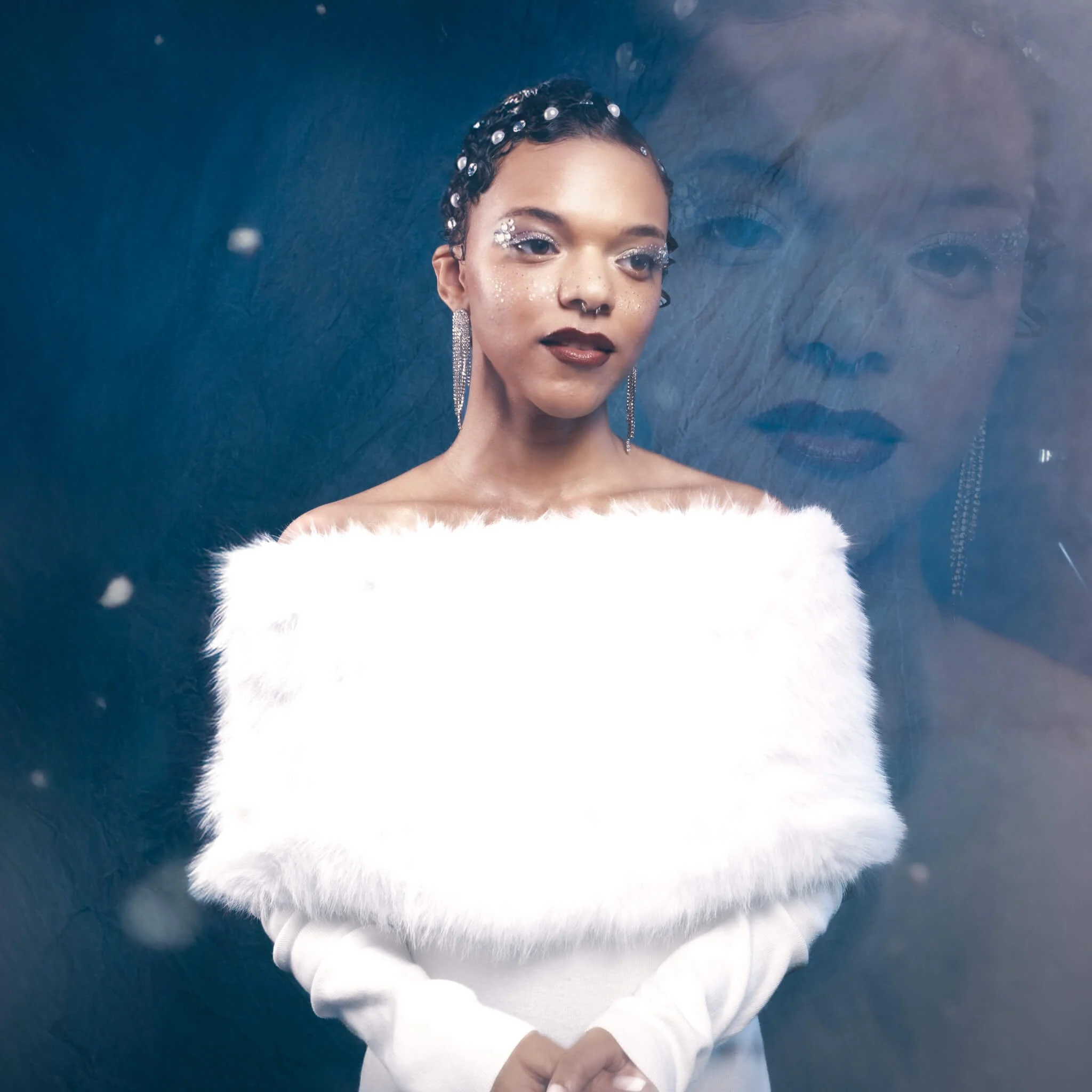 Woman in a white fur wrap and long crystal fringe earrings stands before a deep blue backdrop, her reflection visible in a glass panel beside her, wearing glitter eye makeup, silver freckles, dark red lips, and pearl hair gems