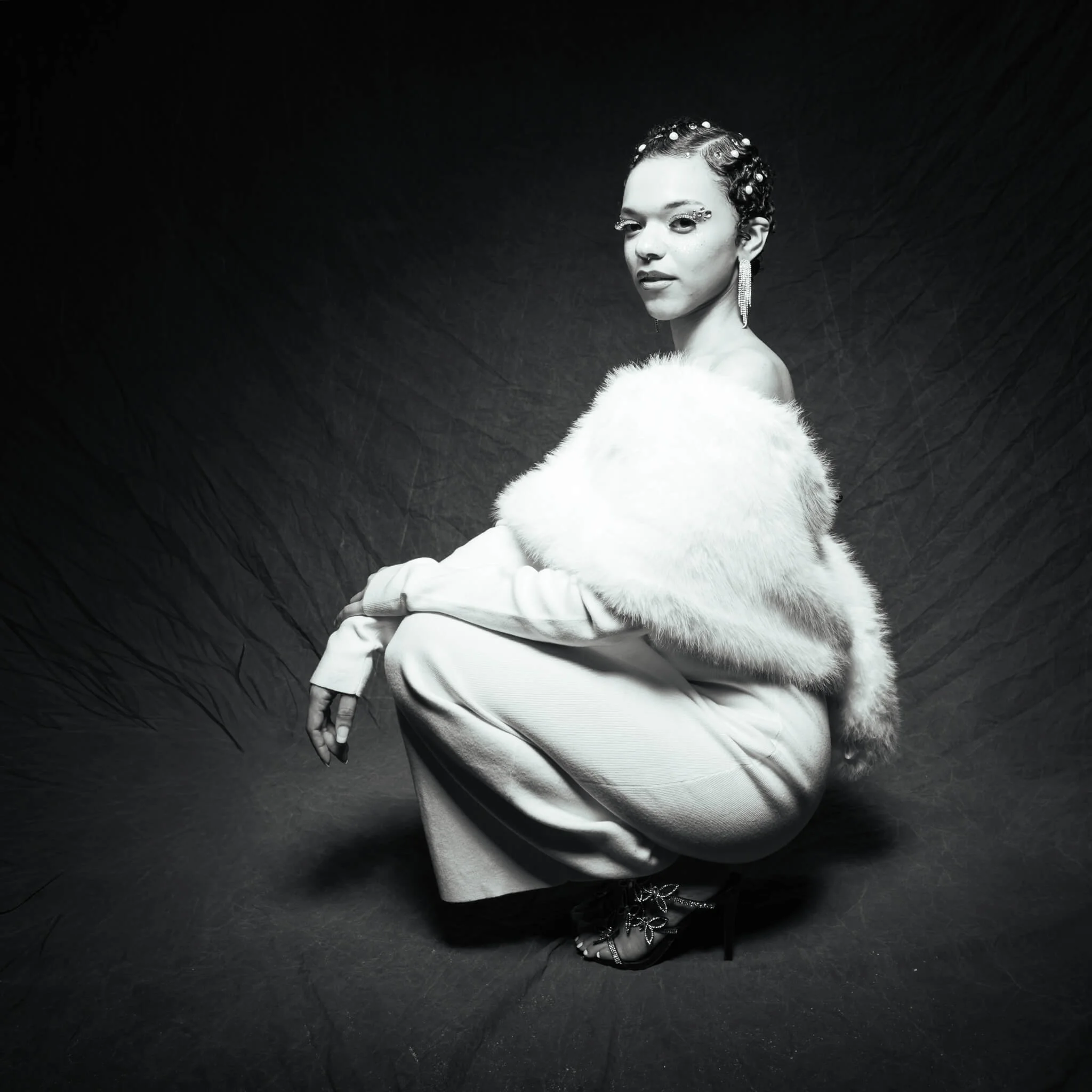 Black and white portrait of a woman crouching low and looking over her shoulder at the camera, wearing a white fur stole, wide-leg trousers, jeweled heeled sandals, crystal fringe earrings, and pearl hair gems against a dark draped backdrop