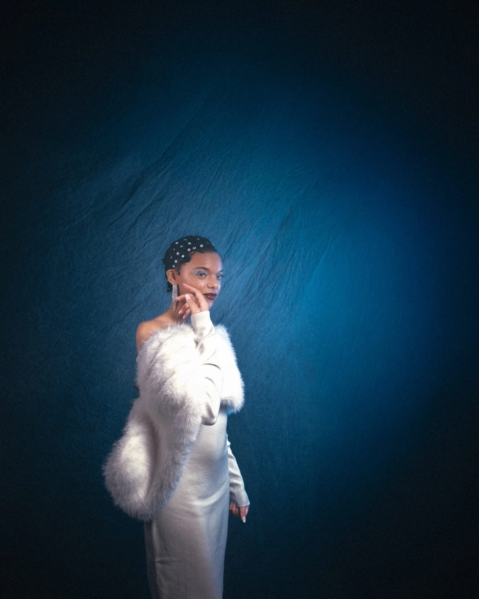 Woman in white off-shoulder dress and white fur stole poses against a deep blue textured backdrop, hand raised to her chin, wearing crystal hair gems and chandelier earrings with glitter eye makeup