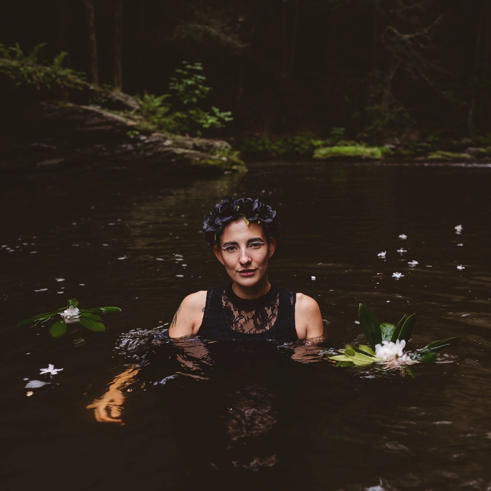 A woman wearing a dark flower crown and black lace dress in a woodland creek, surrounded by floating flowers.