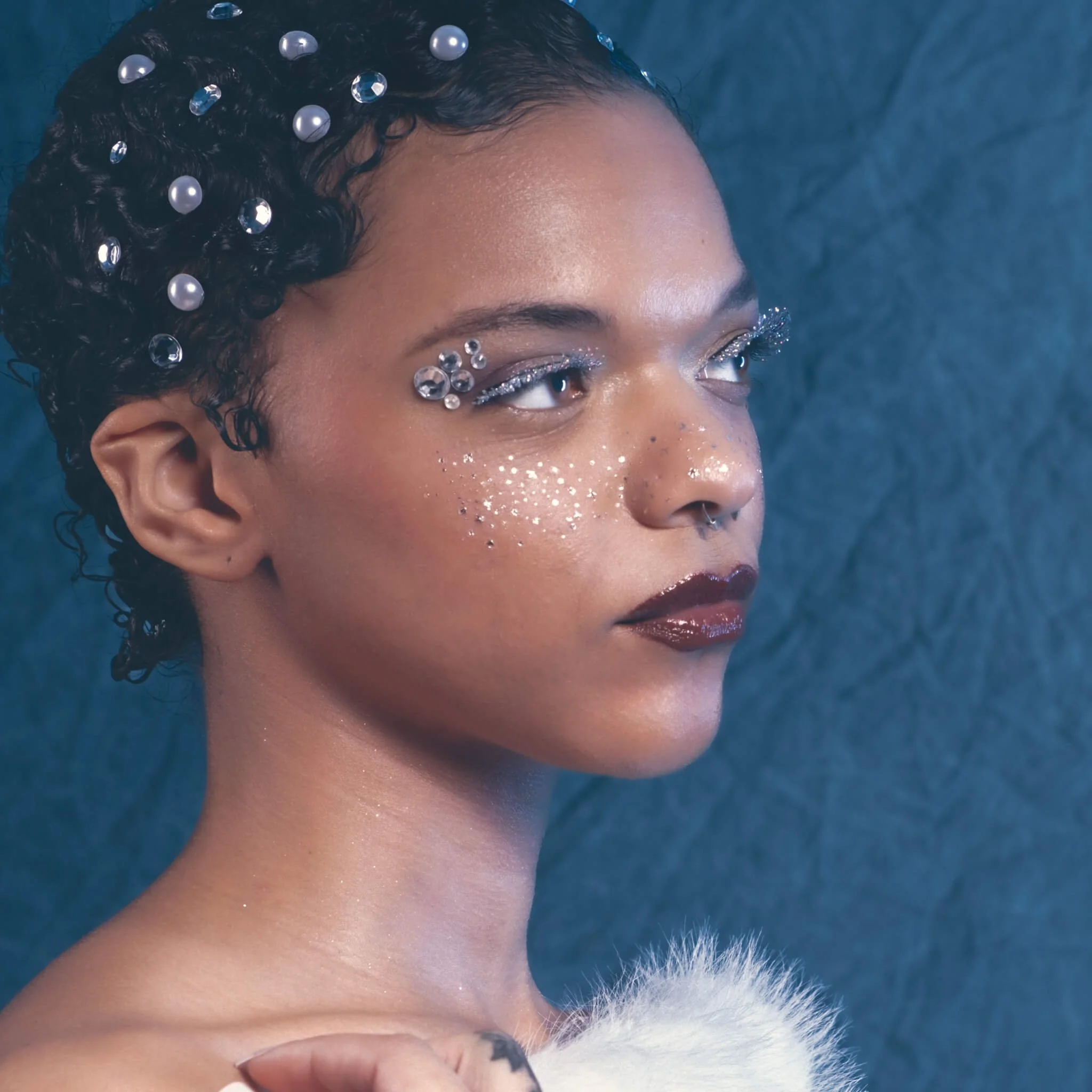 Close-up side profile of a woman with oversized pearl and crystal gems scattered through her hair, jeweled glitter eye makeup, silver glitter freckles, dark red lips, and a white fur stole visible at the shoulder against a deep teal textured backdrop