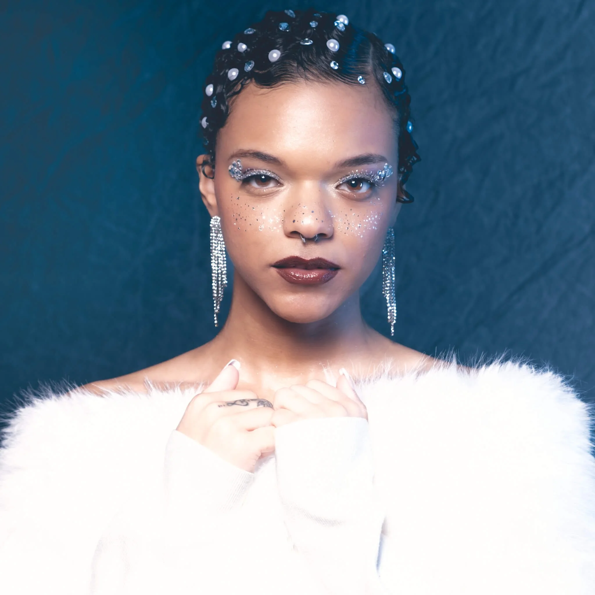 ortrait of a woman gazing directly at the camera with hands clasped at her chest, wearing long crystal fringe earrings, glitter eye makeup, silver freckles, dark red lips, pearl and gem hair gems, and a white fur stole against a deep teal backdrop