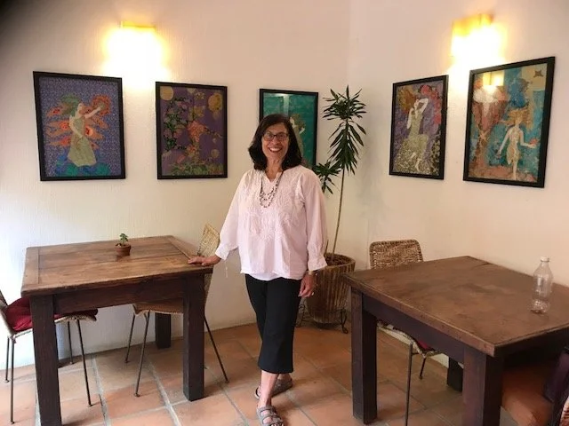 Artist Tanya Sugarman stands in front of a selection of her artwork. She has dark hair and wears a white shirt.