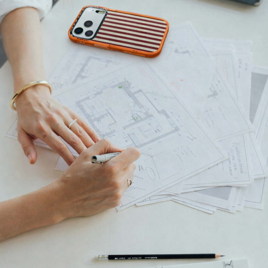 A person is working on architectural plans or blueprints on a white table, with a pen in hand, and a smartphone with a striped case nearby.