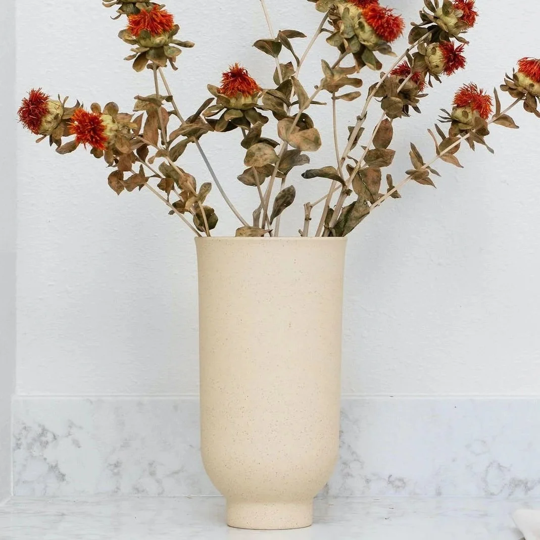 A tall, beige, textured ceramic vase holding dried, reddish-brown and green foliage with small, fuzzy, red flowers.
