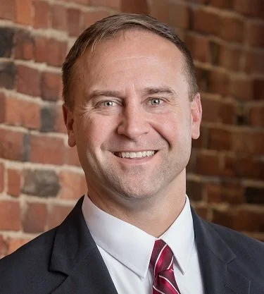 A man in a business suit smiling in front of a brick wall.