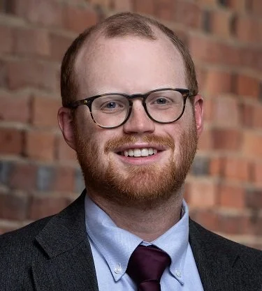 A man with glasses, red hair, and a beard, wearing a suit and tie, smiling in front of a brick wall.