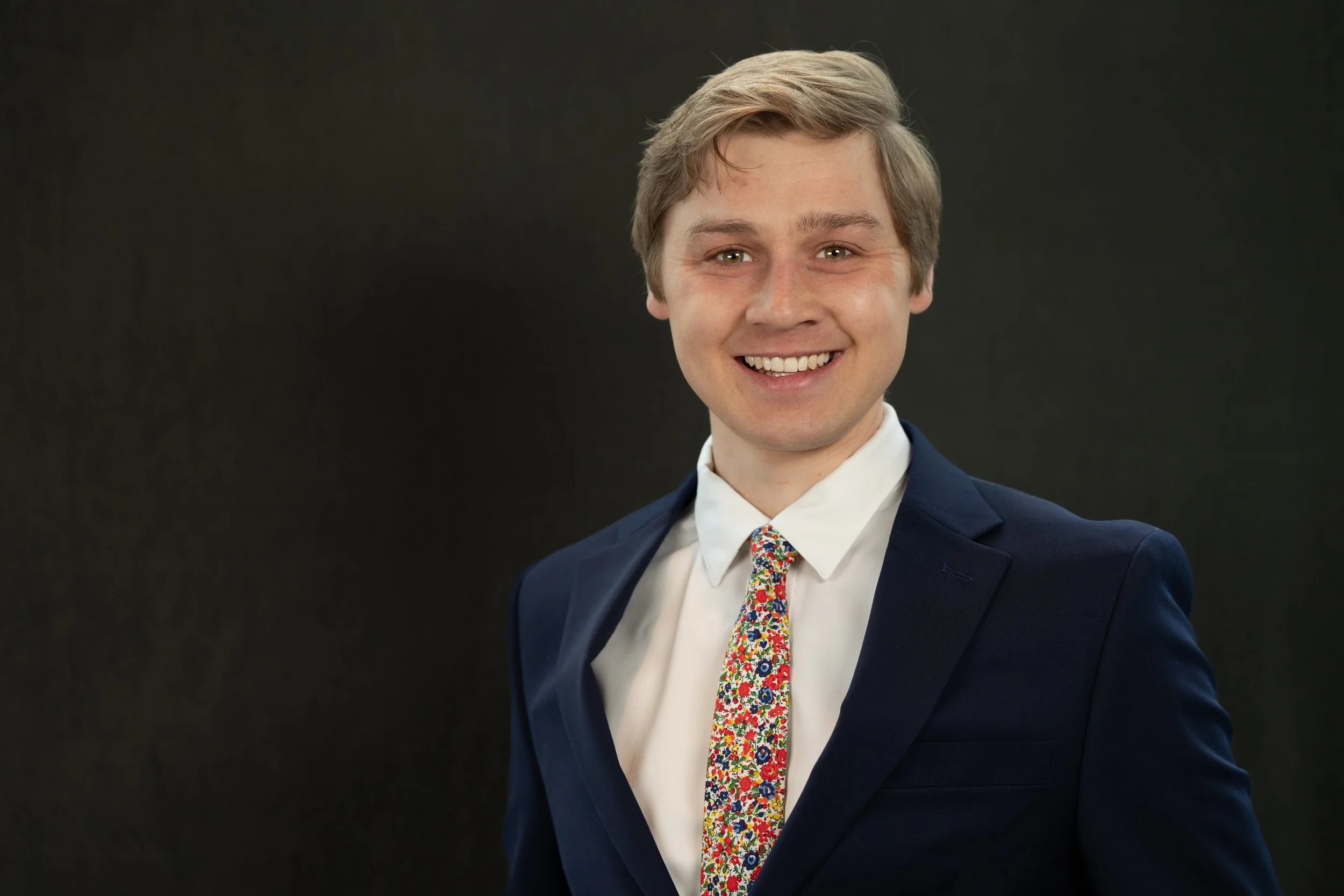Young man smiling outdoors, wearing a dark blazer, white shirt, floral bow tie, and holding a yellow flower. Background is blurred green foliage.