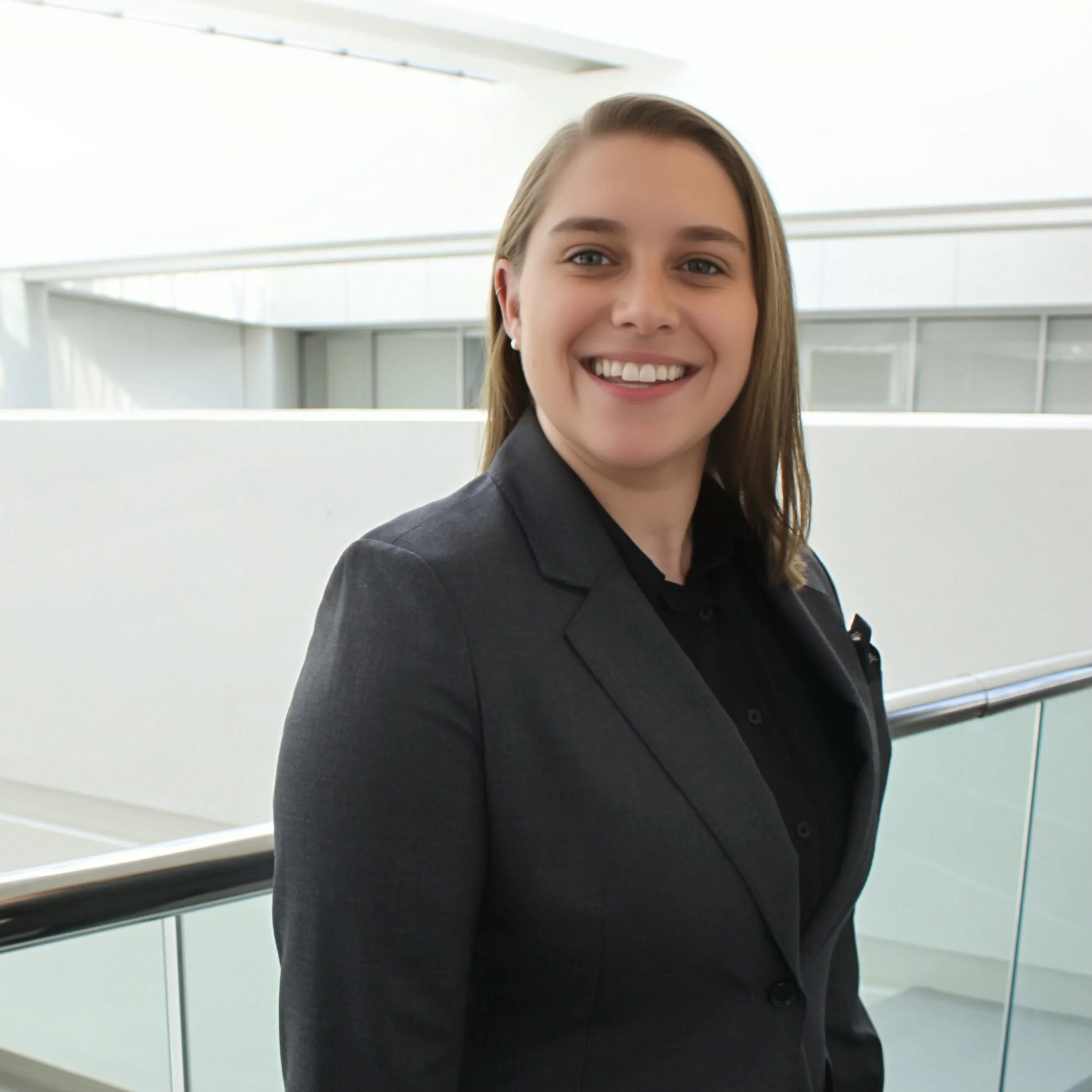 A woman smiling, dressed in a dark blazer and shirt, standing on a modern indoor balcony with glass and metal features.