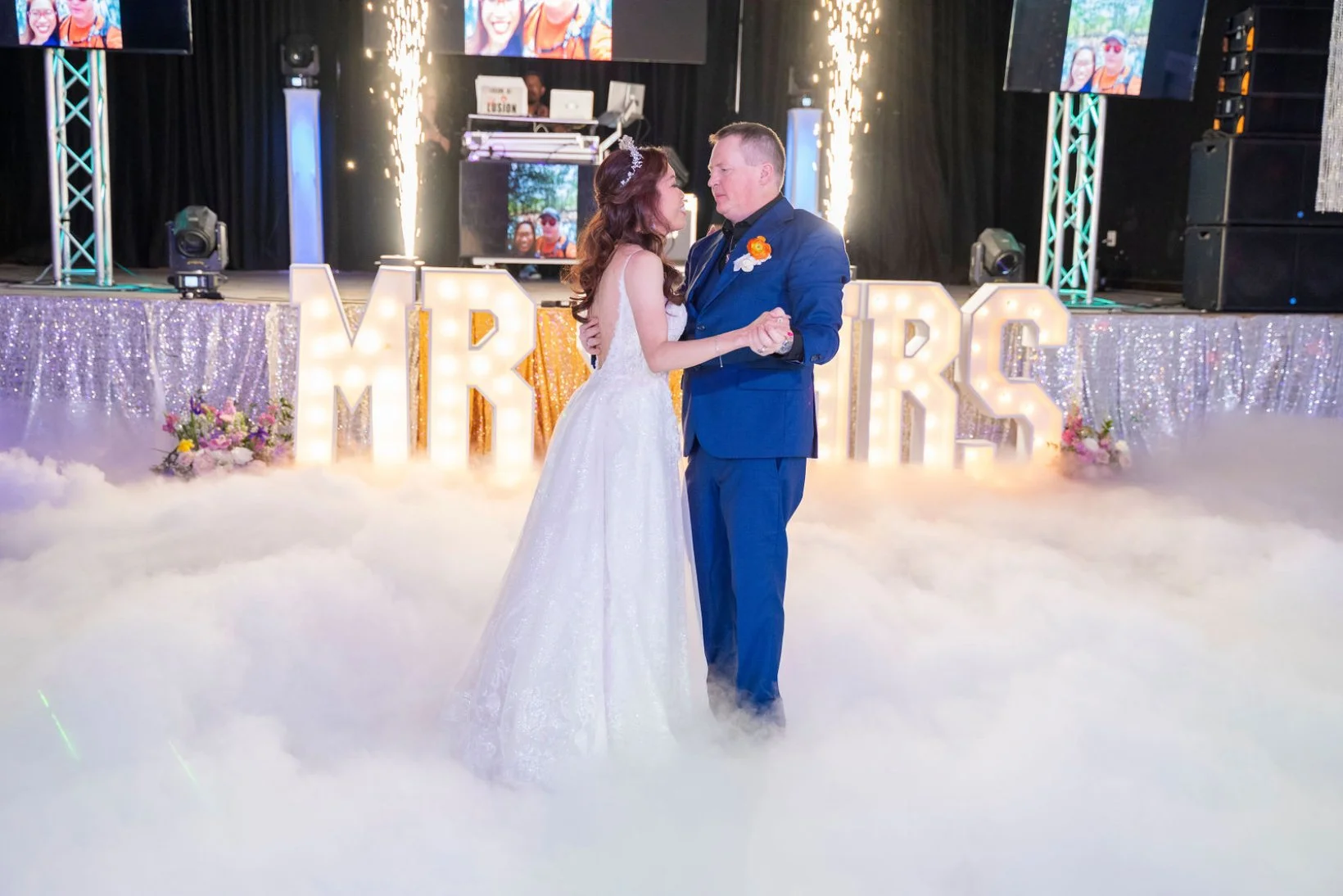 A bride and groom dancing during their wedding reception, with large illuminated letters spelling 'MR & MRS' behind them. The dance floor has a fog-like mist, with sparklers on the stage and decorative flowers around the letters.