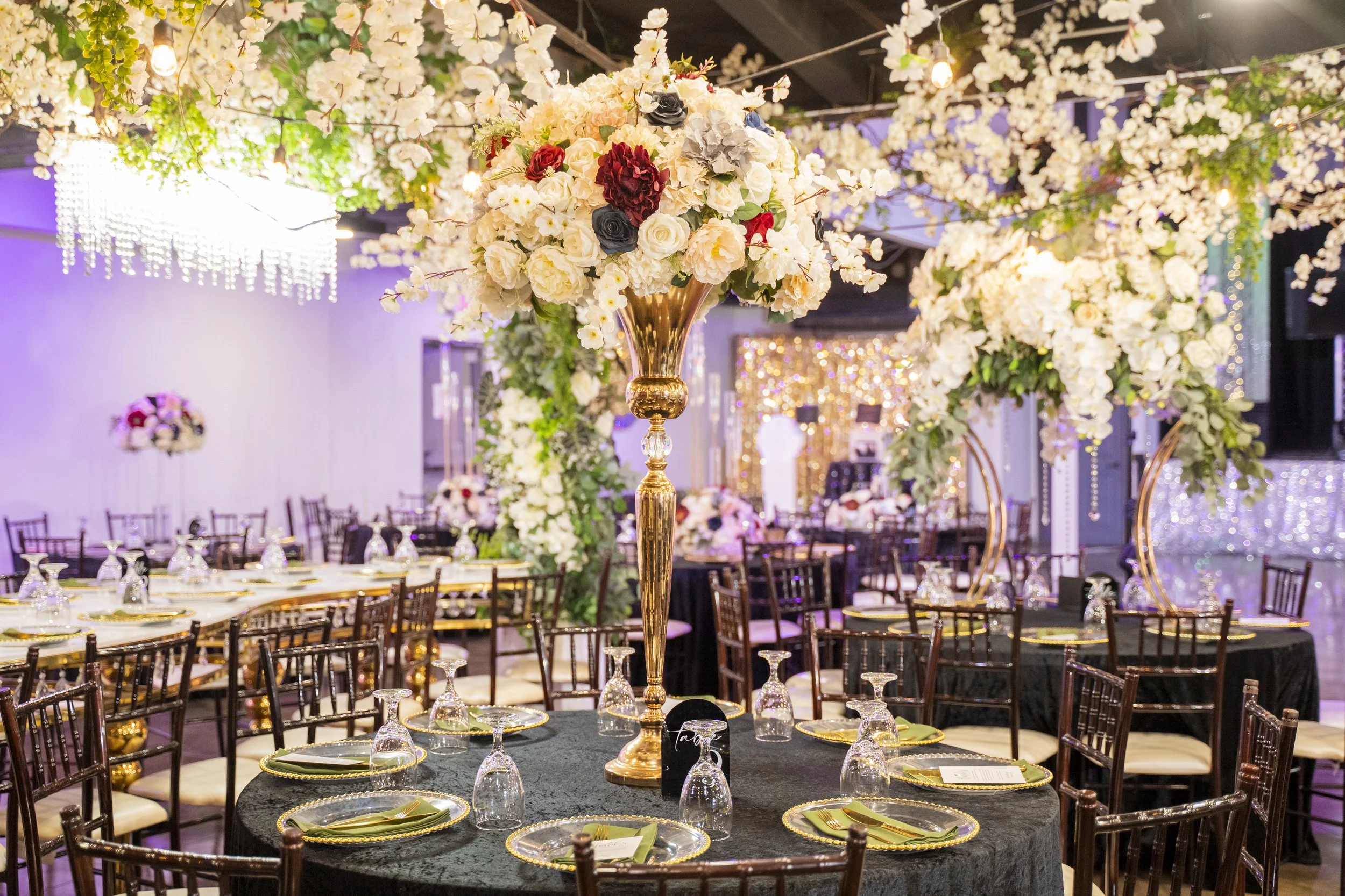 Elegant event venue decorated with tall floral centerpieces of white, red, black, and gray flowers on gold stands, surrounded by tables set with gold-rimmed glass chargers, green napkins, and wine glasses, with string lights and cherry blossom decorations overhead.