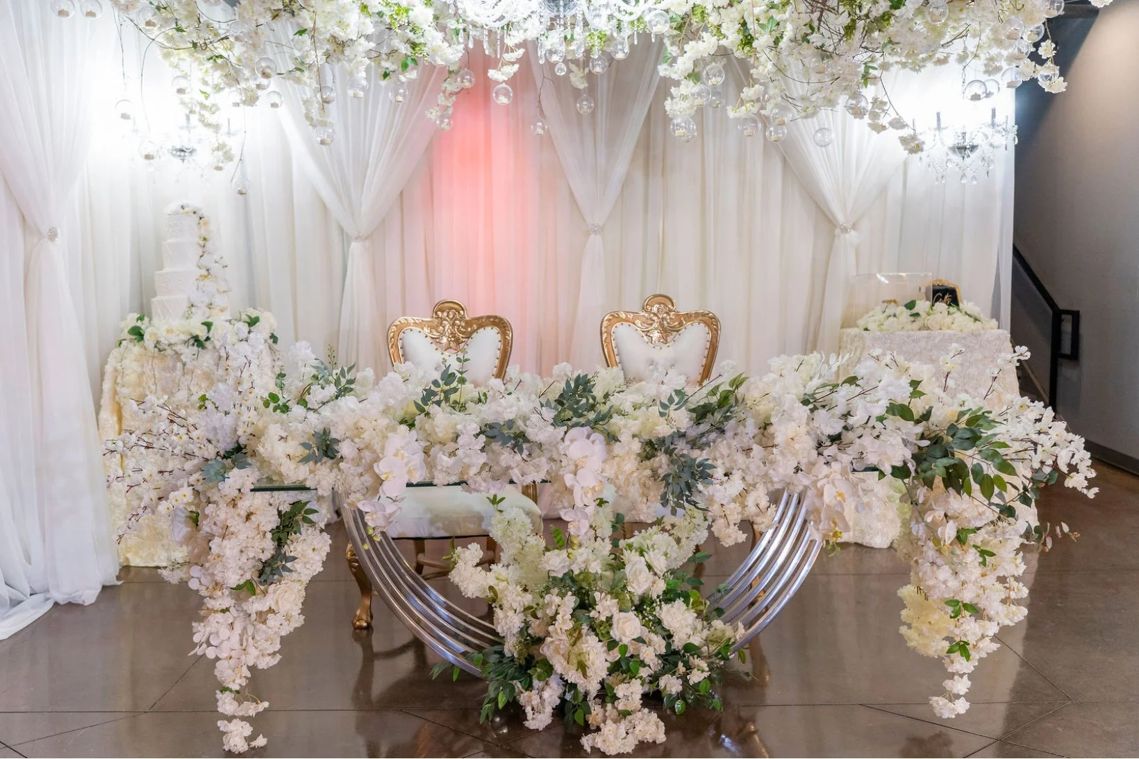 Elegant wedding setup with a floral arch and chairs. Decor includes white flowers, lush greenery, and gold accents, with a backdrop of draped white fabric and chandeliers.