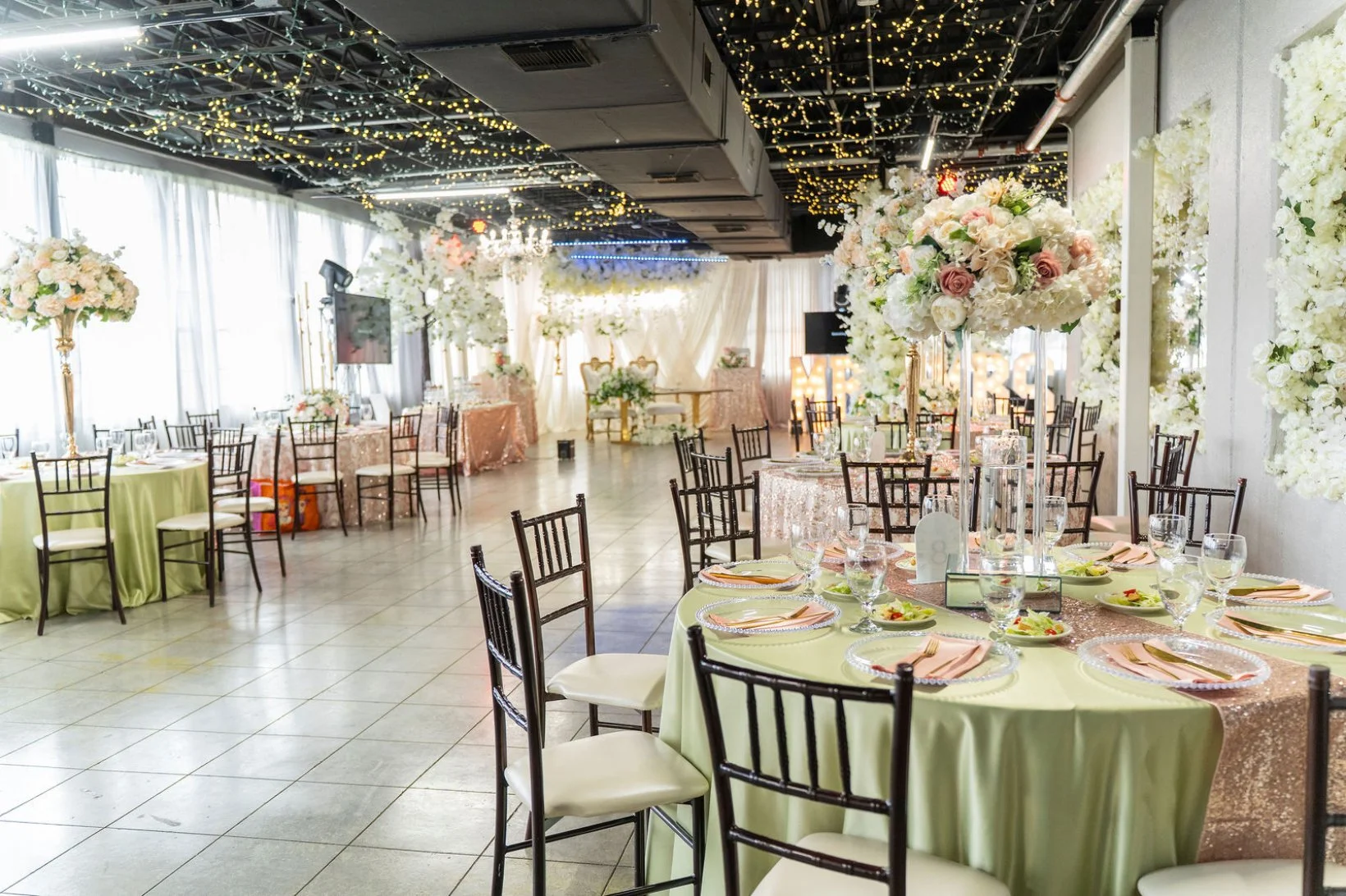 Wedding reception hall decorated with pastel-colored tablecloths, floral centerpieces, and string lights hanging from the ceiling.