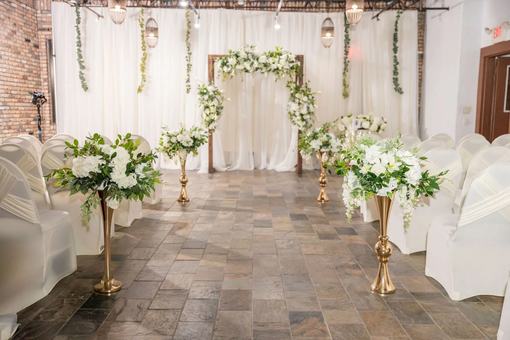 Wedding aisle decorated with white flowers and greenery, with a floral arch at the end, white chairs on either side, and soft lighting in a rustic venue.