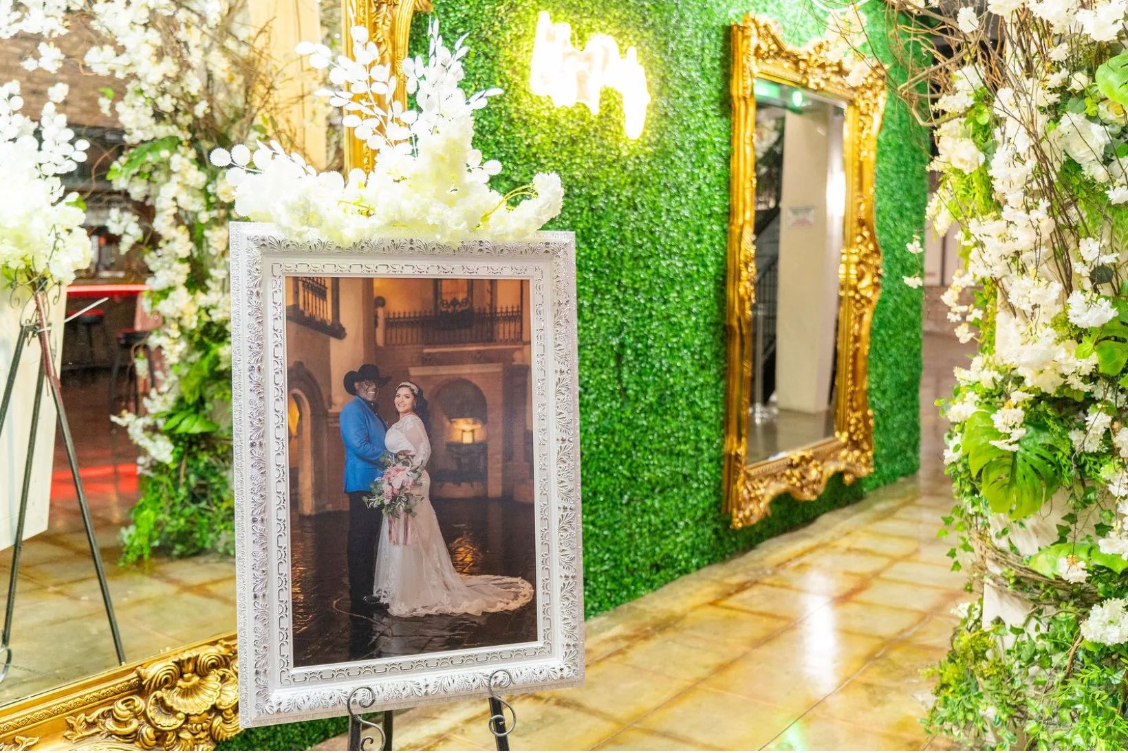 Wedding photo displayed on an easel with a white ornate frame, featuring a couple in wedding attire inside a lavish indoor venue. The background includes a green wall with a logo, floral decorations, and gold-framed mirrors.
