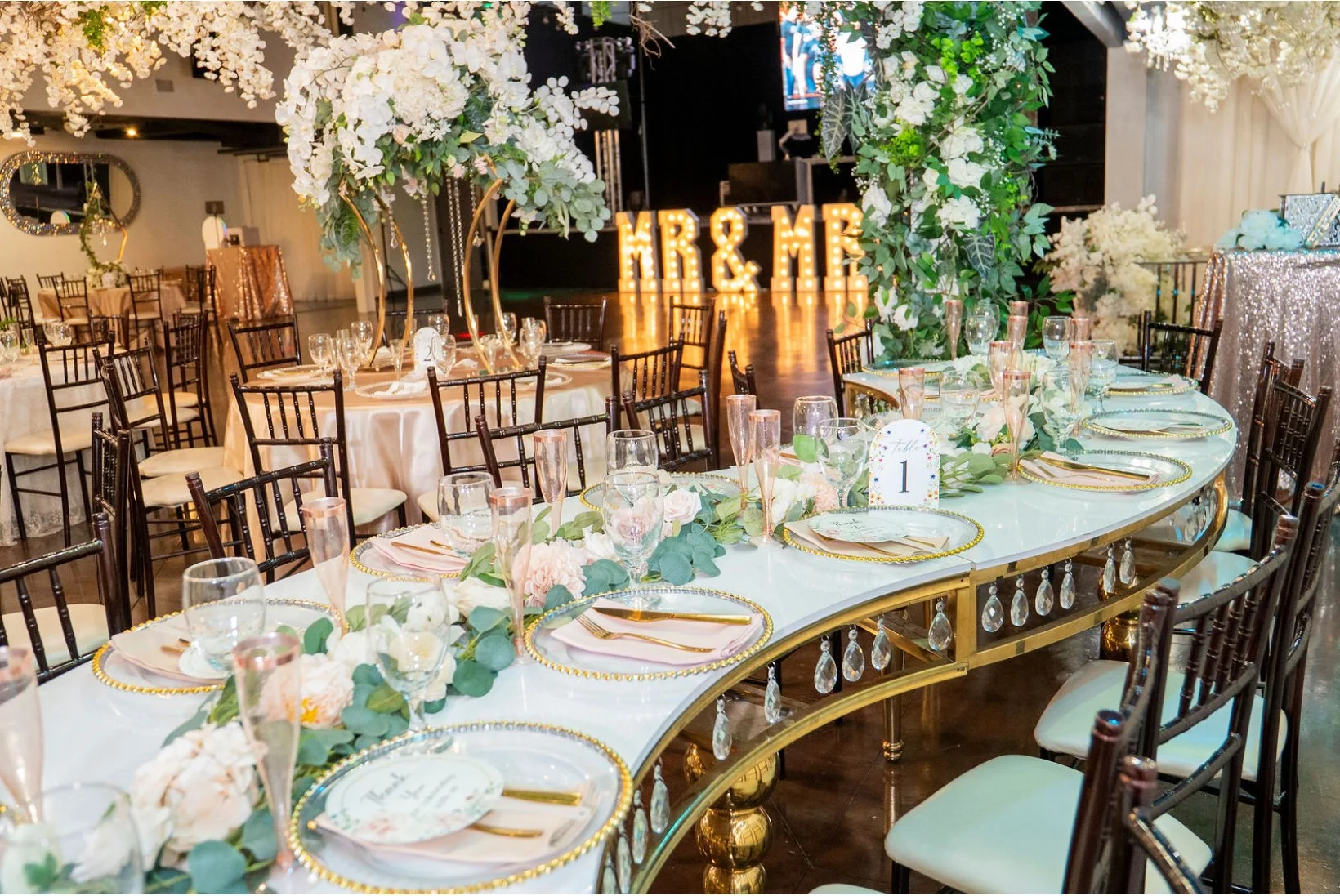 Elegant wedding reception table with floral centerpieces, gold accents, and surrogate pink champagne glasses, in front of large illuminated 'Mr & Mrs' sign.