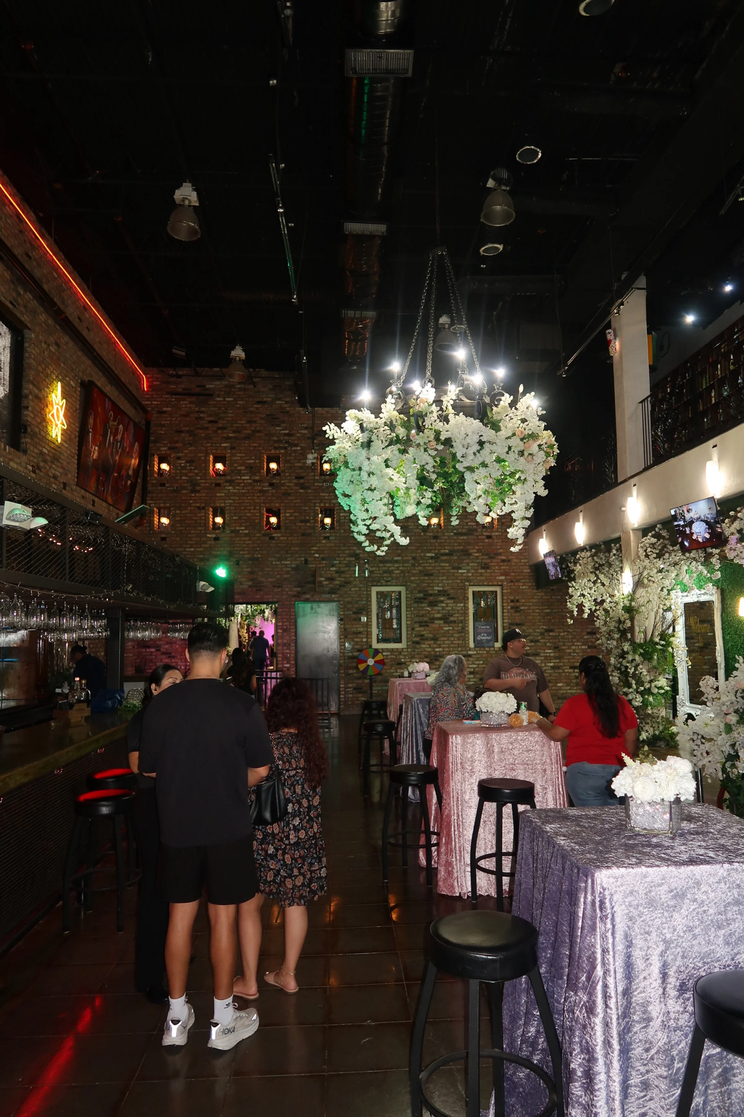 Interior of a decorated event space with floral arrangements, high tables, and guests socializing. There is a large chandelier with white flowers hanging from the ceiling, and a brick wall in the background with small niches and wall decorations.