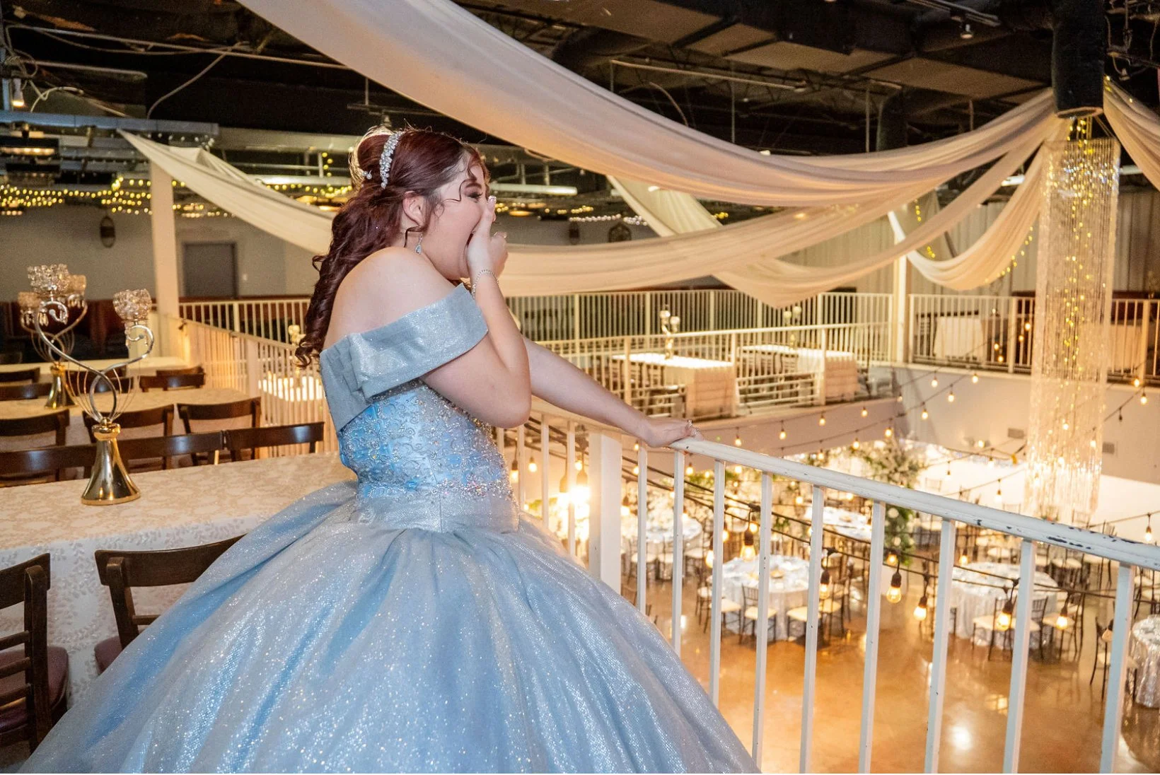 A young woman in a sparkling blue ball gown covering her mouth, standing on a balcony of a decorated event space with draped fabric and string lights.