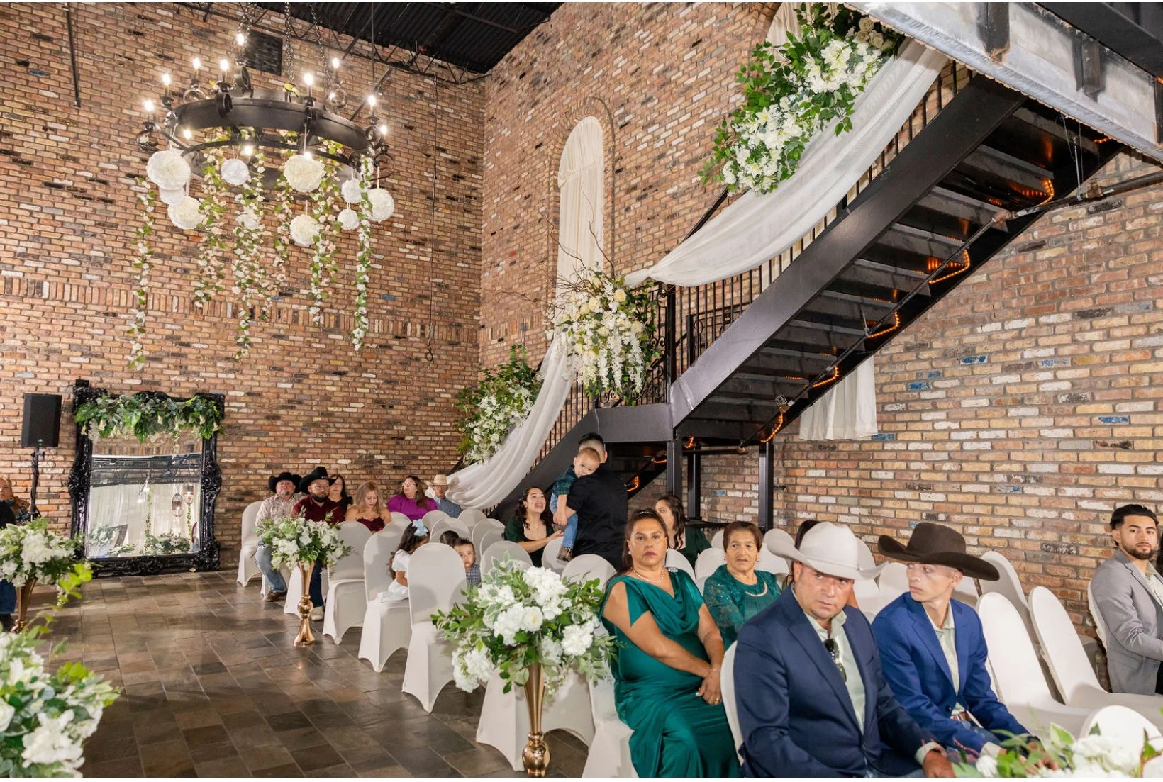 Wedding reception with people sitting on white chairs, decorated in white and green floral arrangements, inside a venue with exposed brick walls, a black staircase, and a chandelier hanging from the ceiling.