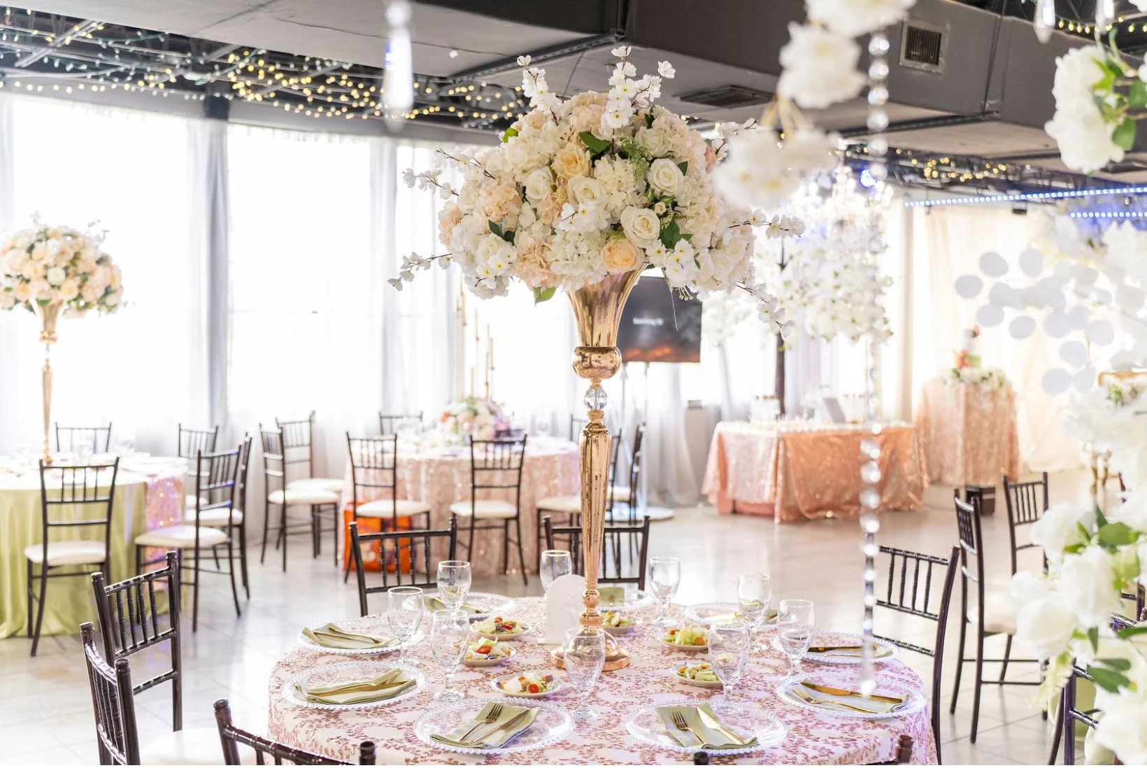 Elegant banquet hall decorated with large flower centerpieces, round tables with pink lace tablecloths, and chairs set for a formal event or wedding reception.