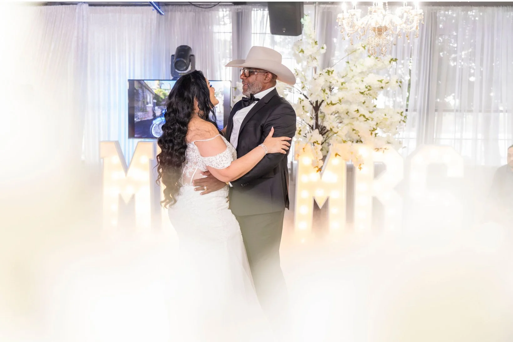 A bride and groom dancing at their wedding reception in a decorated indoor venue with large illuminated letters, white flowers, a chandelier, and a television screen in the background.