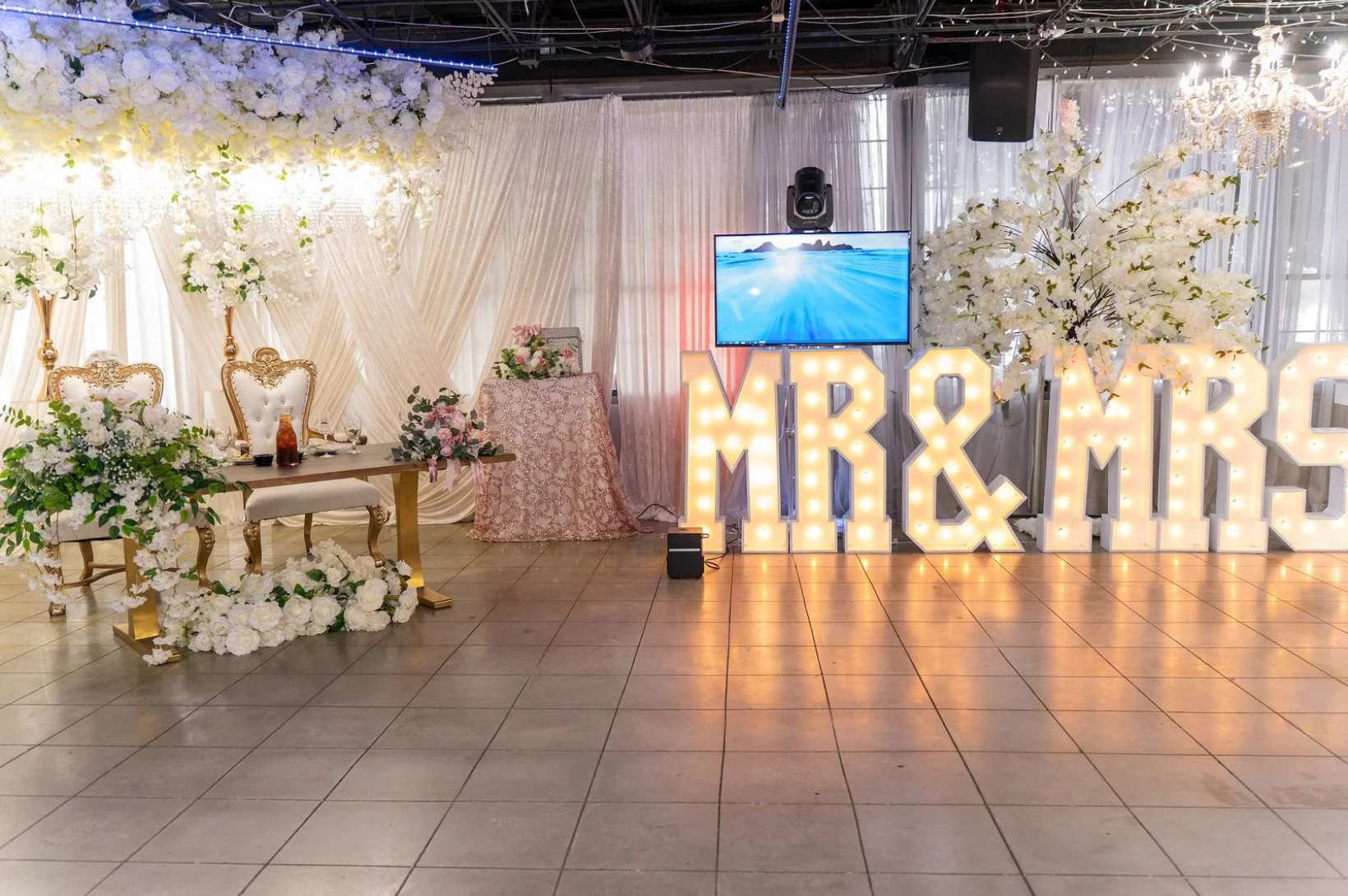 Wedding reception setup with a table, chairs, flowers, large illuminated letters spelling MR & MRS, and a flat screen TV.